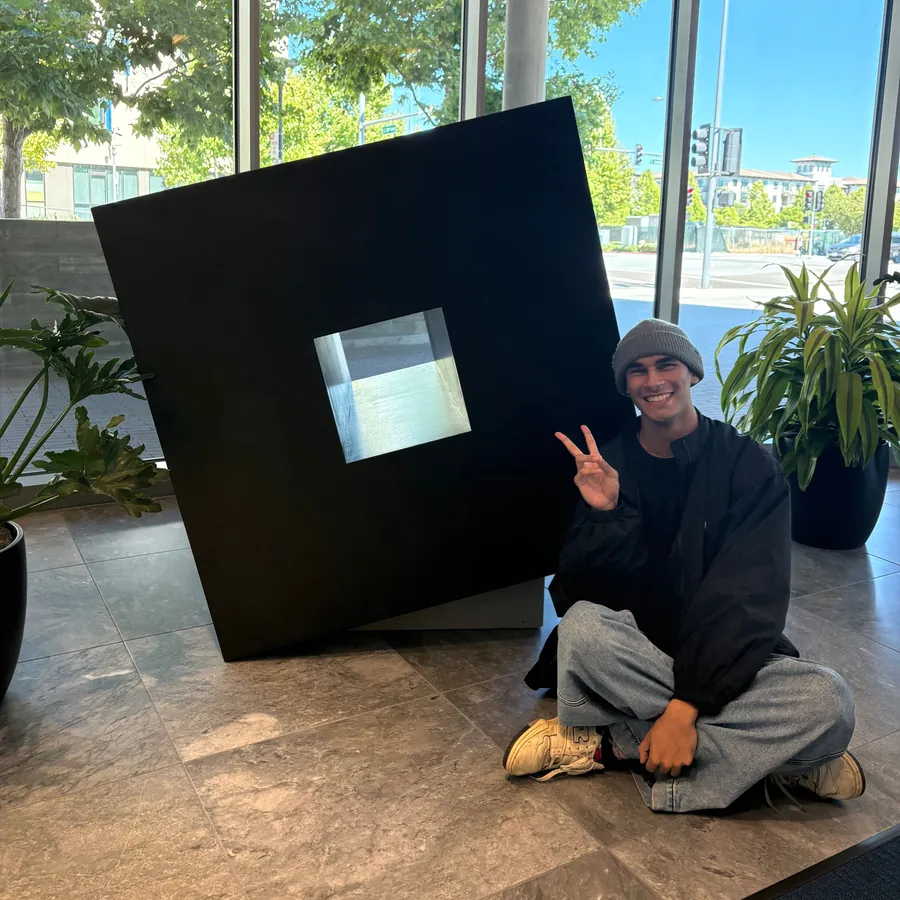 Keyan sitting cross-legged beside the giant tilted Roblox logo in the lobby, making a peace sign